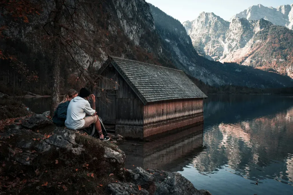 Serene scene of a couple sitting by a lake with rocky mountain views and a wooden cabin reflection.