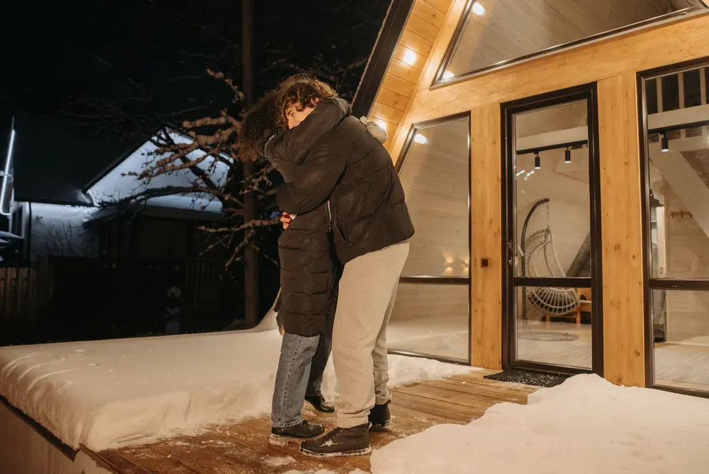 A couple embraces warmly outside a cabin at night, surrounded by snow.