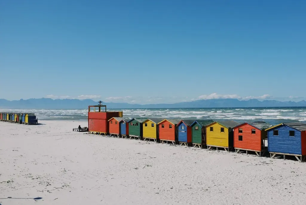 Colorful beach huts on a sandy shore with ocean waves and mountains in the background.