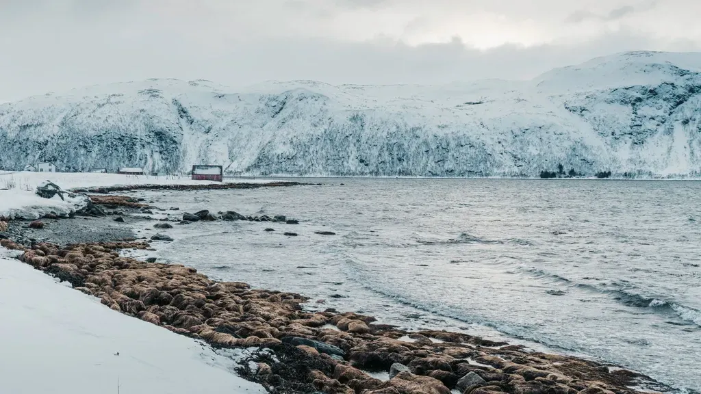 Tranquil winter view of a snowy coastline with a solitary red cabin in the distance against icy mountains.